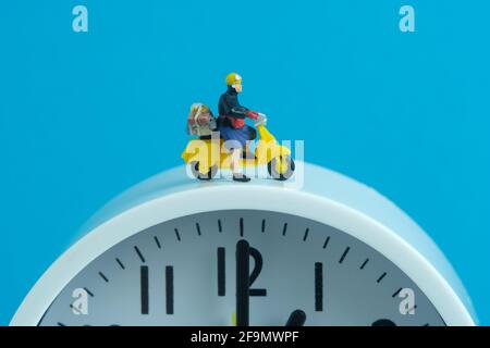 Miniature people toys conceptual photography. Delivery on time service. Postman courier standing above the clock, isolated on blue background. Stock Photo