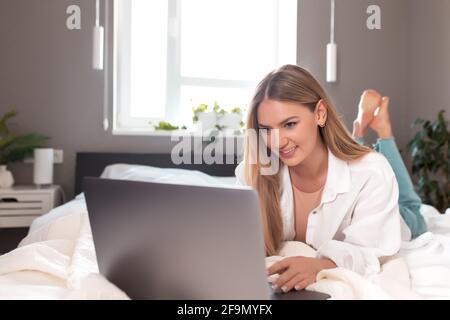 Smiling girl lays on the bed in bedroom and use laptop for remote work Stock Photo