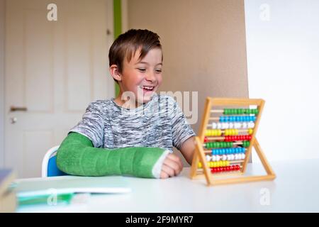 cool boy with green arm cast sits in his room and learns with slide rule with colorful balls made of wood during homeschooling Stock Photo