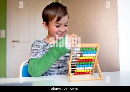 cool boy with green arm cast sits in his room and learns with slide rule with colorful balls made of wood during homeschooling Stock Photo