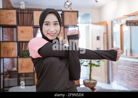 smiling muslim woman in sports clothes stands stretching with one hand holding the other hand when one hand is pulled to the side Stock Photo