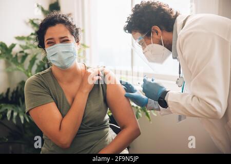Woman getting scared while receiving a covid vaccine at home. Male healthcare professional giving vaccine to female. Stock Photo