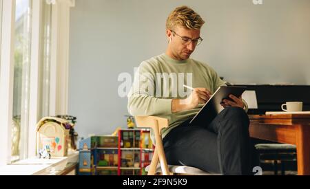 Hardworking man drawing sketches on his tablet computer. Illustrator using graphic tablet for making new designs while sitting in a chair at home. Stock Photo