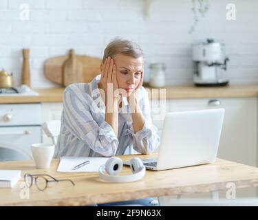 Stressed mature woman in casual wear sitting in modern kitchen, holding head in hands and looking on laptop screen while working or studying online from home. Senior people and technologies concept Stock Photo