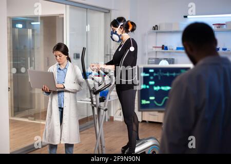 Team of professional doctors measuring health conditions of woman athlete in science sport laboratory. Physician using laptop controling EKG data, muscle endurance, heart rate psychological resistance. Stock Photo