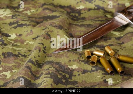 A knife blade and some random casings on camouflage fabric. Knife without sheath and Group of pistol and rifle empty casings lying on military uniform Stock Photo