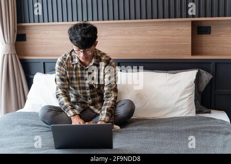 work from home, A Checkered shirt man who wearing eye glasses sit on the bed while using computer,full body. Stock Photo
