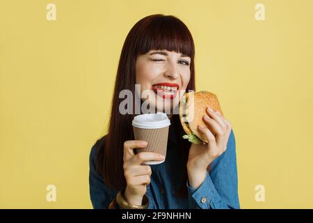 Close up of a cheerful young attractive woman grimacing at camera, showing tongue, holding tasty burger and take away coffee and enjoying her lunch time. Isolated on yellow studio backgroud Stock Photo