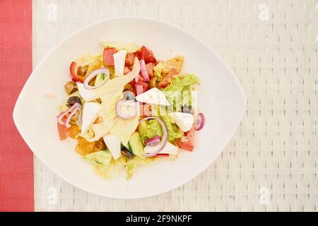 Vegetarian salad in ceramic plate on white raffia table. Delicious greek salad with red onion, lettuce, cucumber, olives, cheese and peppers. Oil sauce with sesame. Healthy food. Stock Photo
