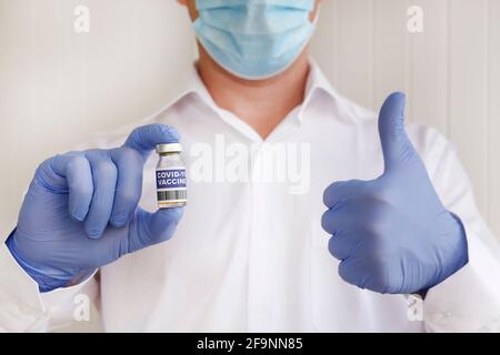 Macro shot of a doctor wearing face mask and latex gloves demonstrating the new successful covid-19 vaccine in vial and showing thumbs up gesture. Clo