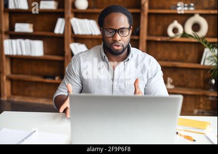 Concentrated young African-American office worker in glasses looking at the webcam of the laptop and gesturing, confident black man using compter app for video connection with colleagues Stock Photo