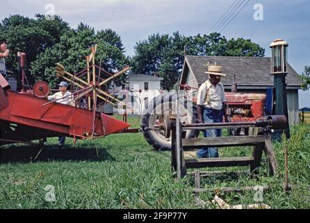 A tractor is being re-fuelled from a bowser on a farm in the USA in the early 1950s. Two white farm workers converse by the harvesting equipment (left), whilst a black farm worker walks in front of the tractor. This image is from an old American amateur Kodak colour transparency – a vintage 1950s photograph. Stock Photo