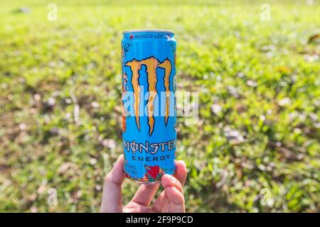 A hand holding the base of Monster Energy drink against a green field background. Stock Photo