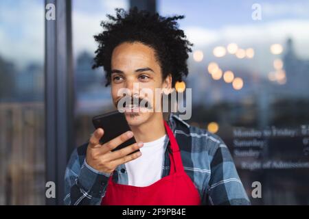 Happy mixed race male barista with moustache leaning in the doorway of cafe talking on smartphone. independent small business in a city. Stock Photo