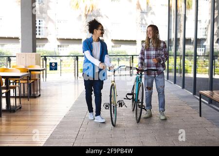 Two happy mixed race male friends wheeling bicycles in the street and talking. green urban lifestyle, out and about in the city. Stock Photo