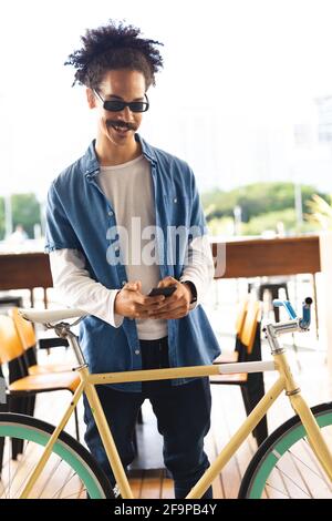 Happy mixed race man with moustache standing in street with bicycle using smartphone. digital nomad, out and about in the city. Stock Photo