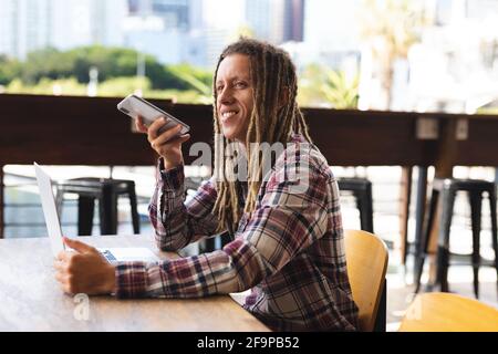 Smiling mixed race man with dreadlocks sitting at table outside cafe talking on smartphone. digital nomad, out and about in the city. Stock Photo