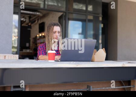 Mixed race man with dreadlocks sitting at table outside cafe using laptop. digital nomad, out and about in the city. Stock Photo