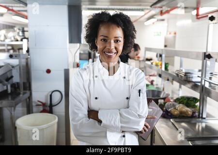 Portrait of smiling mixed race female professional chef with colleagues in background. working in a busy restaurant kitchen. Stock Photo