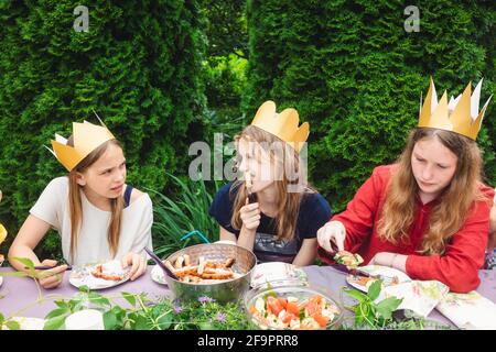 Children wearng paper crowns sitting by  decorated table eating grilled sausages celebrating birthday party in a green garden Stock Photo