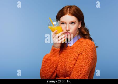 bored redhead woman in sweater holding fresh orange juice in plastic cup isolated on blue Stock Photo