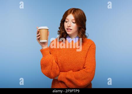 redhead woman in orange sweater holding coffee to go while biting lips isolated on blue Stock Photo