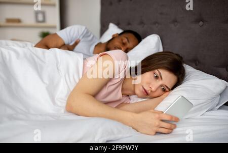 Curious young woman checking her sleeping husband's smartphone in bed at home Stock Photo
