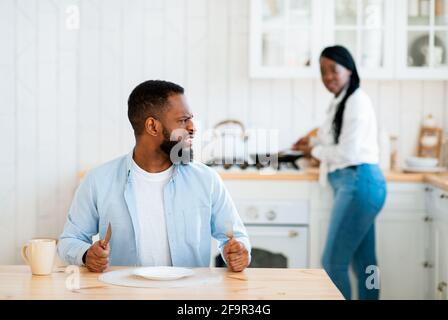 Impatient Hungry Black Husband Waiting For Food, Sitting At Table In Kitchen Stock Photo