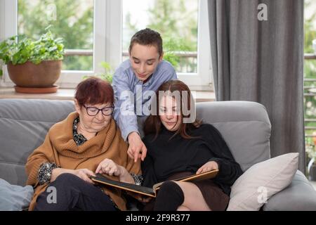 Multigenerational family, Smiling Family Watching into Photo Album on Sofa. Grandmother and grandchildren watching old photo album at home. Stock Photo