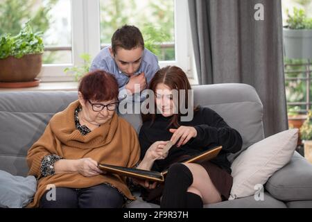 Multigenerational family, Smiling Family Watching into Photo Album on Sofa. Grandmother and grandchildren watching old photo album at home. Stock Photo