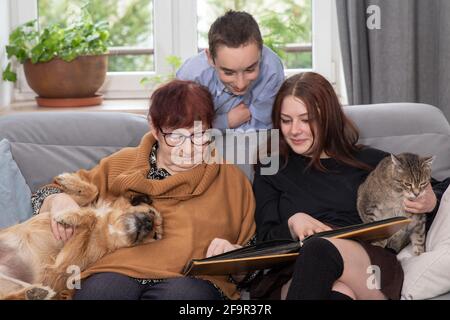 Multigenerational family, Smiling Family Watching into Photo Album on Sofa. Grandmother and grandchildren watching old photo album at home. Stock Photo