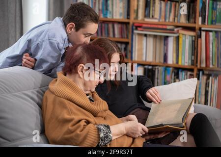 Multigenerational family, Smiling Family Watching into Photo Album on Sofa. Grandmother and grandchildren watching old photo album at home. Stock Photo