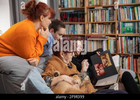 Multigenerational family, Smiling Family Watching into Photo Album on Sofa. Grandmother and grandchildren watching old photo album at home. Stock Photo