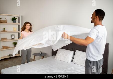 Domestic chores. Millennial multinational couple in love making bed at home, having fun while doing housework together Stock Photo