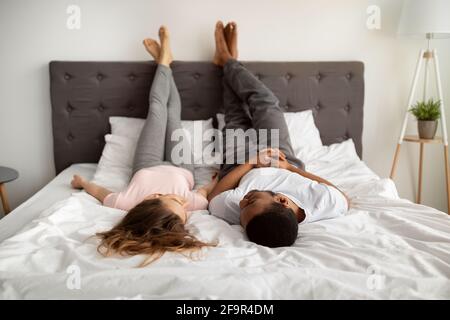 Millennial interracial couple in love lying on bed at home, raising legs up, talking to each other Stock Photo