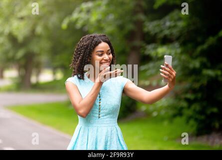 Happy black girl talking to her boyfriend online, sending air kiss on mobile phone at park Stock Photo