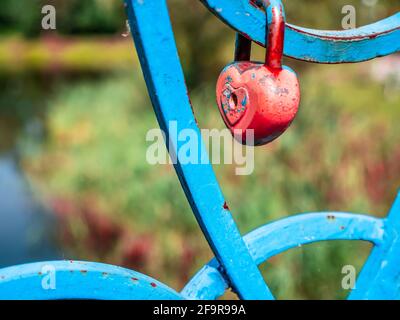 An old wedding lock in heart shape with soft edges and red peeling paint on a blue painted bridge fence under bright sunshine. Stock Photo
