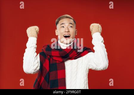 happy young man in a sweater and scarves on a red background holds his hands in a fist Stock Photo