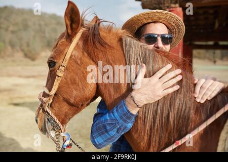 young man hat and beard hugging his horse in the field Stock Photo