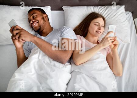 Gadget addiction and relationship problems. Millennial interracial couple with smartphones lying in bed, top view Stock Photo