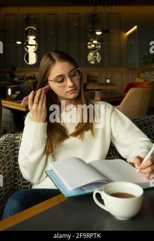 Woman in glasses listening to the audio message, writing note in cafe Stock Photo