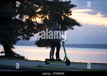 A young couple share a scooter along the Alki Trail as the sun sets over the Olympic Mountains in Seattle, on Monday, April 19, 2021. Stock Photo