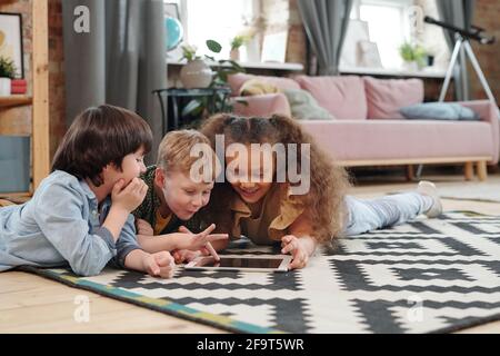 Group of happy children lying on the floor and playing in digital tablet together in the room at home Stock Photo
