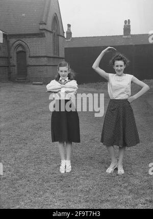 1956, historical, outside in the grounds of a church, two  girls in their outfits, showing the moves they will perform in the traditional May Queen carnival, England, UK. An ancient festival of Spring and celebrations, in many villages, May Day involved the crowning of a May Queen and dancing around a Maypole, activities that have taken place in England for centuries. Selected from the girls of the area, The May Queen wearing a crown, would start the procession of floats and dancing. In the industrialised North of England, the Church Sunday Schools often led the organisation of the day. Stock Photo