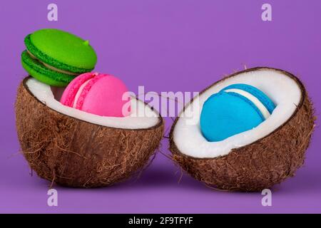 Looking for the summer! Vibrant pink, green, yellow and orange French macarons and fresh coconut fruit half, on light purple background. Stock Photo