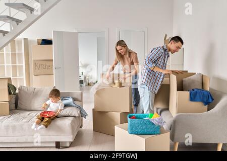 Happy family new home owners with child unpacking cardboard boxes on moving day. Stock Photo