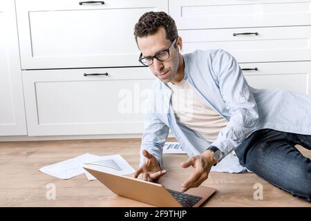 Concentrated businessman lays on floor at home, using laptop, developing new project. Thoughtful stylish male employee or student is working or Stock Photo