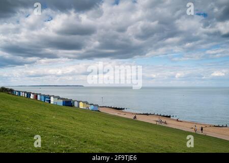 Whitstable, UK - April 4th 2021 Pretty colourful beach huts along a seafront with groynes. There is grass in the foreground. People are walking and cy Stock Photo