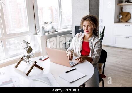 Stylish female freelancer is using laptop, working on startup, thinking about ideas. Attractive young business woman sitting at the desk in home Stock Photo