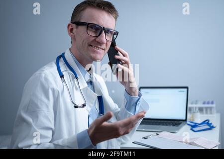 Smiling doctor having phone call at desk. Physicianr talking with his patient in office clinic sitting at table with laptop. German doctor in white Stock Photo